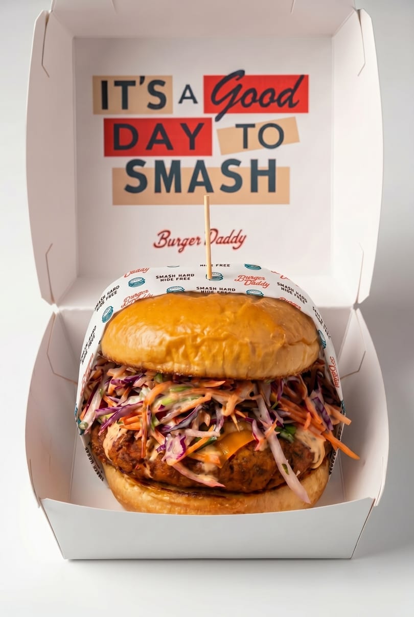 A Burger Daddy burger with slaw and sauce in an open takeaway box with "IT'S A Good DAY TO SMASH" printed inside.