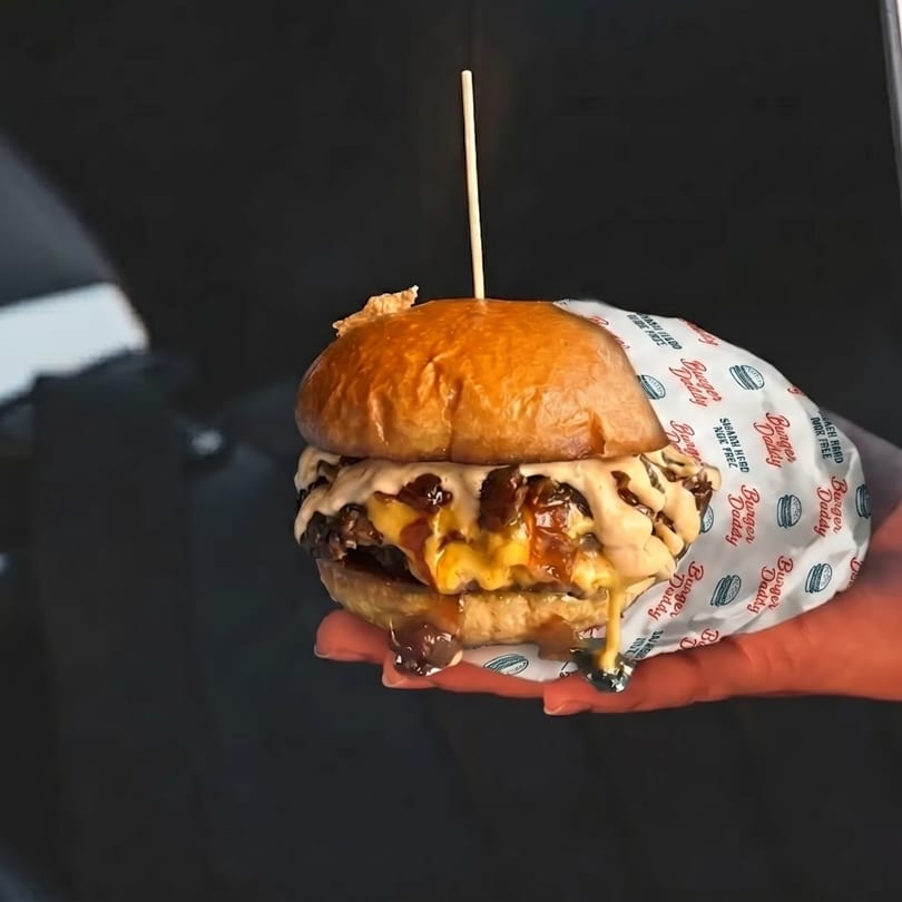 A hand holds a Burger Daddy double cheeseburger with melted cheese and sauce, wrapped in branded paper.